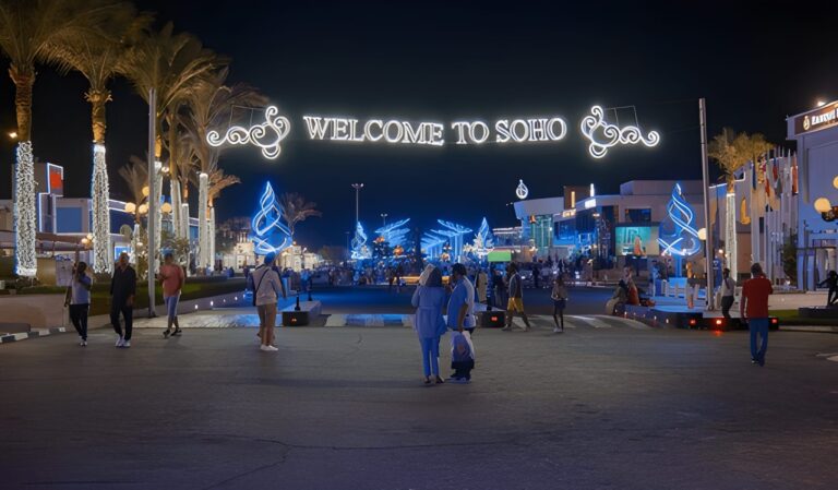 Night view of SoHo in New York, beautifully lit with blue and white lights and people walking under the “Welcome to SoHo” sign.